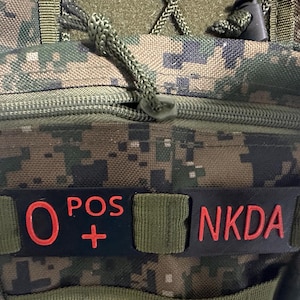 May include: A camouflage patterned tactical pouch featuring a black rectangular patch with the red text O POS + and NKDA. The pouch includes a zippered compartment, olive green webbing straps, and a green cord pull tab for the zipper closure.