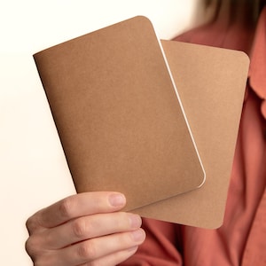 May include: Two rectangular notebooks with plain, light brown kraft paper covers and rounded corners. The notebooks are held together, showing a simple, unadorned design with a smooth texture and a clean, minimalist appearance for writing or sketching.