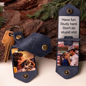 May include: Navy blue leather keychain with a photo insert. The keychain has a snap closure and a gold-colored key ring. The photo insert displays a picture of a person with a cat. The text "ALEX" is embossed on the keychain.