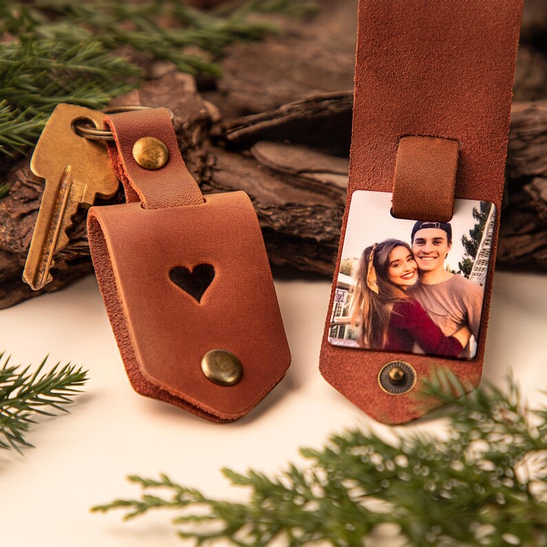 May include: Two brown leather keychains. One has a heart cutout and a brass button. The other has a photo attached and a brass button. Both keychains are attached to a key and are on a wooden surface.