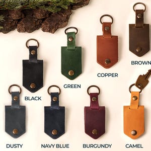 May include: A collection of leather keychains in various colors, including black, green, copper, brown, dusty, navy blue, burgundy, and camel. Each keychain features a metal ring and a snap closure. The word "BROWN" is printed on one of the keychains.