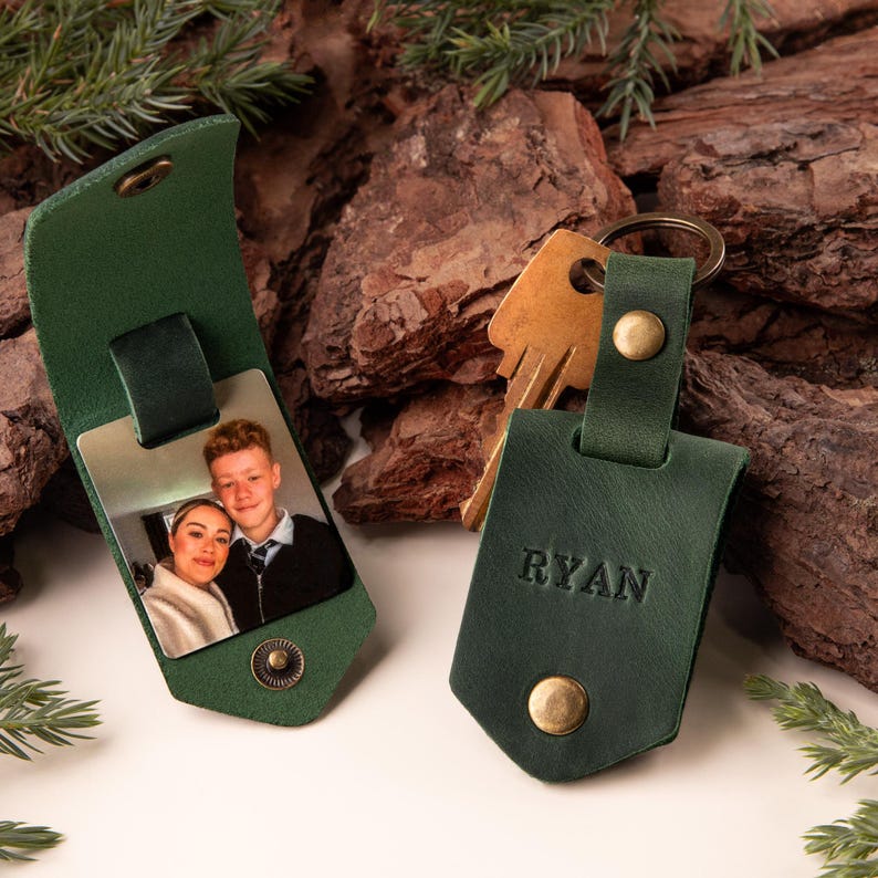 May include: Green leather keychain with a gold snap and the name "RYAN" embossed on it. The keychain is next to a matching leather case with a photo. The background includes wood and greenery.
