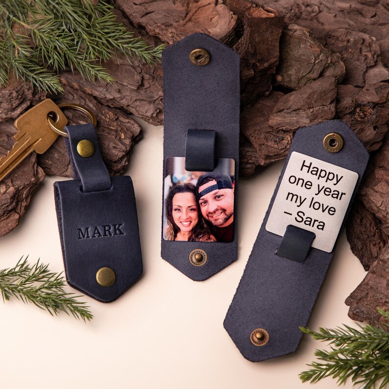 May include: Three navy blue leather keychains. One keychain has the name "MARK" embossed. Another displays a photo of a couple. The third keychain has the text "Happy one year my love - Sara". Each keychain has a brass snap.