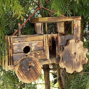 May include: A rustic, wooden birdhouse shaped like a tractor, with a round entry hole and a chimney. The birdhouse is made of weathered wood and has a dark, burnt finish. It hangs from a tree branch.