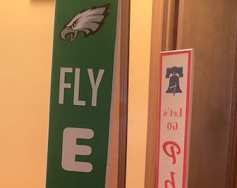May include: Green and white sign with the words "FLY EAGLE" and a picture of a bald eagle. A red and white sign with the words "Let's go Phils" and a picture of the Liberty Bell.