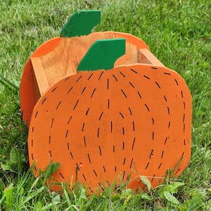 May include: Orange pumpkin-shaped wooden planter with green painted leaves and black lines. The planter is sitting on green grass.