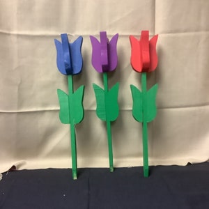 May include: Three wooden tulips painted in blue, purple, and red. The tulips have green stems and leaves.