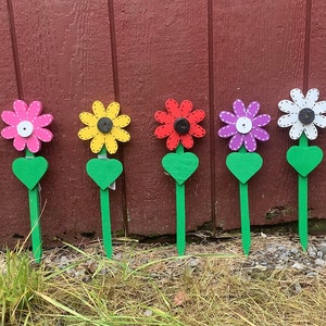 May include: Eight wooden flower stakes with heart-shaped leaves. The flowers are painted in various colours, including orange, pink, yellow, red, purple, white, and blue. Each flower has a black button in the centre.