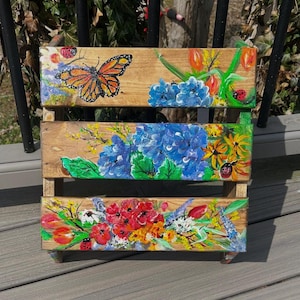 May include: A wooden, three-tiered planter box with hand-painted floral and insect designs. The top tier features a butterfly and blue flowers. The middle tier has blue hydrangeas, and the bottom tier displays red flowers. The box is painted with bright colors.
