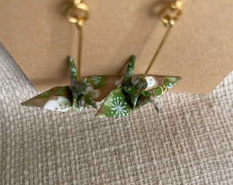 May include: A pair of gold earrings with green and white origami cranes. The cranes have a floral pattern and are hanging from gold hooks.