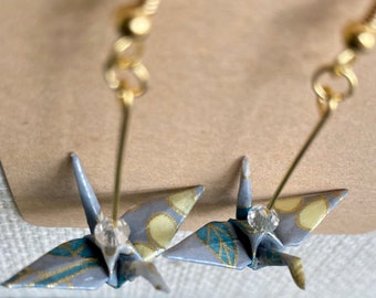 May include: A pair of gold origami crane earrings with a blue and gold patterned paper design and clear crystal accents.