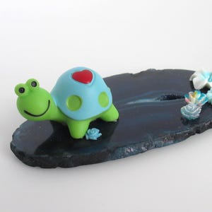 May include: A green turtle figurine with a light blue shell featuring a red heart and green spots, positioned on a dark blue, polished agate slice. The agate is decorated with several small, colorful, multi-petaled flower embellishments.