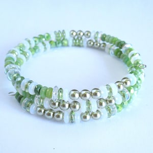 May include: A multi-strand wrap bracelet featuring a repeating pattern of metallic gold beads, opaque white disc beads, and translucent light green faceted beads. The design consists of three circular layers that overlap to form a continuous coil.