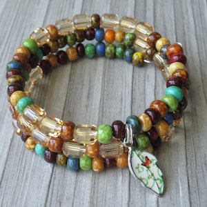 May include: A multi-strand wrap bracelet featuring a mix of round beads in shades of green, brown, blue, and yellow, interspersed with clear faceted cylindrical beads. A white leaf-shaped charm with a small orange butterfly graphic hangs from it.