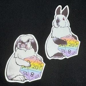 May include: Two die-cut stickers featuring white rabbits with dark ears and paws. Each rabbit holds a multi-colored, rainbow-gradient twenty-sided die, or d20, with black numbers clearly visible on the faces, including the number 20 at the center.