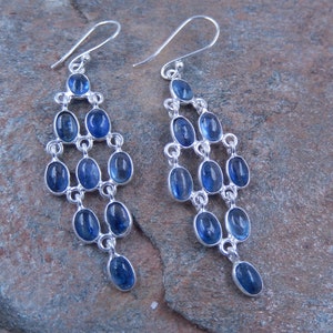 May include: A pair of chandelier earrings with a silver-tone metal setting. Each earring features multiple oval-shaped, deep blue gemstones arranged in a cascading design. The earrings have a hook closure.