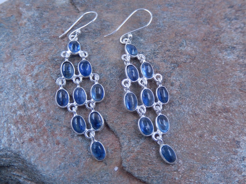 May include: A pair of chandelier earrings with a silver-tone metal setting. Each earring features multiple oval-shaped, deep blue gemstones arranged in a cascading design. The earrings have a hook closure.