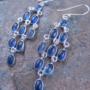 May include: Silver chandelier earrings with multiple tiers of oval, blue gemstones. The earrings have a cascading design, with each tier connected by silver links. The gemstones are a vibrant blue color, and the earrings have a hook closure.