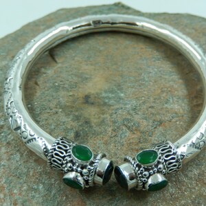 Op de afbeelding: Een zilveren armband met een getextureerd, gehamerd oppervlak langs de band. De twee open uiteinden zijn afgewerkt met sierlijk zilverfiligraan en kraalwerk, elk bezet met drie kleine, ovaalvormige, levendig groene edelstenen.