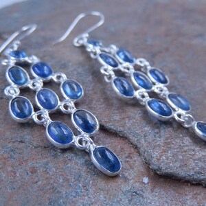 May include: Elegant chandelier earrings with a cascade design. Each earring features multiple oval-shaped, deep blue gemstones set in a silver-toned metal. The gemstones are arranged in a tiered, flowing pattern, creating a sophisticated and eye-catching accessory.
