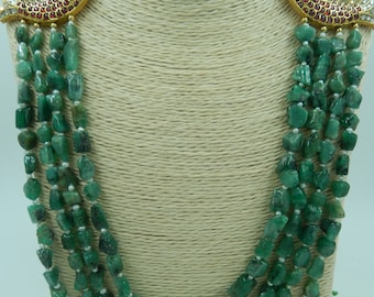 Puede incluir: Un collar de cuatro hileras de piedras preciosas de color verde esmeralda con colgantes de peces de oro y detalles de rubí rojo. El collar se muestra en un maniquí marrón.