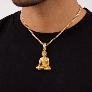 Puede incluir: Un collar con colgante de Buda dorado. El colgante representa una figura de Buda sentado. La cadena es de estilo eslabones dorados. La persona de la imagen lleva una camiseta negra.
