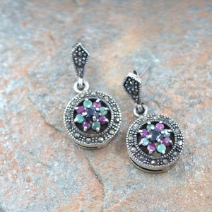 May include: A pair of silver dangle earrings with a round, floral design. The earrings are adorned with a cluster of small, colorful gemstones, including green, pink, and black.