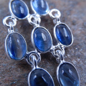 May include: A silver-toned pendant featuring multiple oval-shaped, deep blue gemstones. The stones are set in individual bezels and connected by delicate links, creating a layered design. The gemstones have a polished, reflective surface.