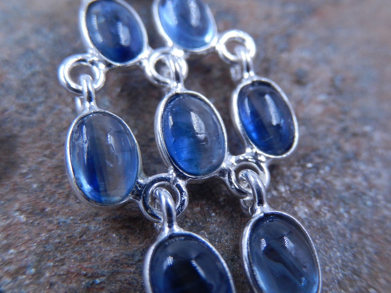 May include: A silver-toned pendant featuring multiple oval-shaped, deep blue gemstones. The stones are set in individual bezels and connected by delicate links, creating a layered design. The gemstones have a polished, reflective surface.