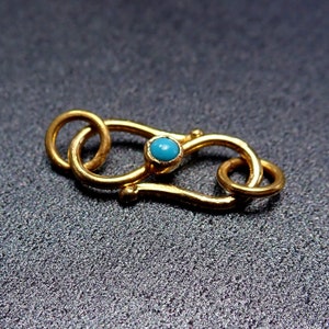 Könnte beinhalten: Ein goldfarbener Verschluss mit einem türkisfarbenen Stein in der Mitte. Der Verschluss hat ein geschwungenes Design mit zwei Schlaufen an einem Ende und einem Haken am anderen. Der türkisfarbene Stein ist rund und in eine goldene Lünette gefasst.