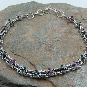 Puede incluir: Un brazalete de cadena de plata con piedras preciosas verdes y rosas alternadas. El brazalete tiene un cierre de langosta.