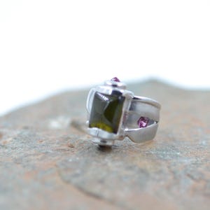 May include: A silver ring with a rectangular, olive-green gemstone at the center. The ring features two small, round, pink gemstones and a unique, geometric design. The ring is set on a textured, gray surface.