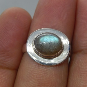 May include: A silver ring featuring an oval-shaped labradorite gemstone. The stone has a gray base with iridescent blue and green flashes. The ring's setting is a simple, polished silver band.