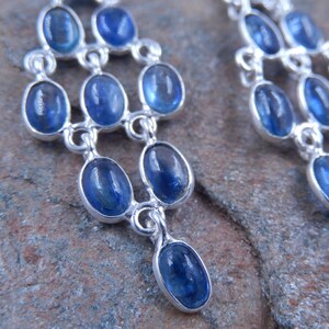 May include: Close-up of a pair of silver chandelier earrings with oval, deep blue gemstones. The earrings have a tiered design, with each tier featuring multiple gemstones. The gemstones are set in a silver metal frame.