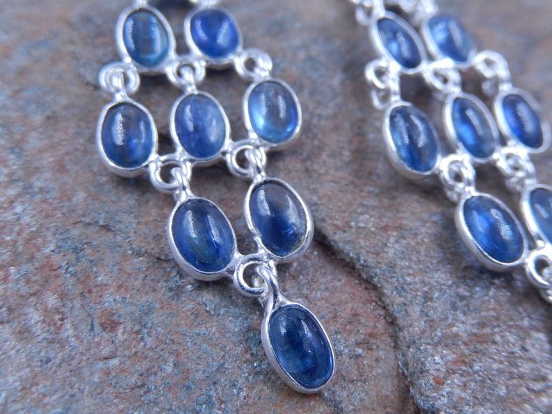 May include: Close-up of a pair of silver chandelier earrings with oval, deep blue gemstones. The earrings have a tiered design, with each tier featuring multiple gemstones. The gemstones are set in a silver metal frame.