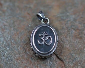 May include: Oval silver pendant featuring the Om symbol in the center. The pendant has a dark background and a decorative border. The bail is also silver. The pendant is displayed on a textured, gray-brown surface.