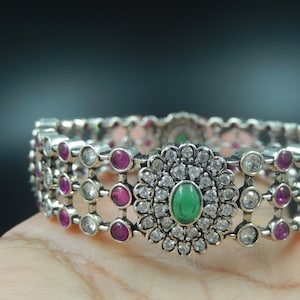 May include: A silver bangle bracelet with a green stone surrounded by white stones in a floral design. The bracelet is decorated with pink stones.