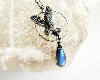 May include: A silver owl pendant necklace with a teardrop-shaped blue labradorite gemstone. The owl is depicted in flight and is surrounded by a silver circle.