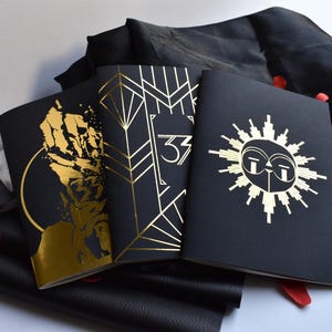 Peut inclure: Plusieurs carnets noirs avec des motifs dorés, dont des motifs géométriques, un soleil et une silhouette. Le chiffre "33" est présent sur plusieurs articles. Des pétales rouges et du tissu noir complètent la composition.