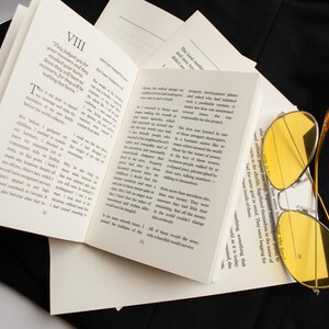 May include: An open book with printed text and yellow-tinted sunglasses resting on the pages. The book is surrounded by black fabric. The glasses have a gold-colored frame.