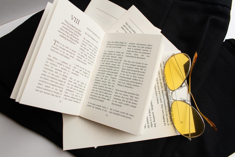 May include: An open book with printed text and yellow-tinted sunglasses resting on the pages. The book is surrounded by black fabric. The glasses have a gold-colored frame.