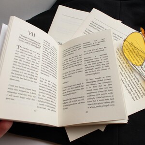 May include: An open book with text on its pages, surrounded by loose sheets of paper and yellow-tinted sunglasses. The book is held open by a hand, with the text visible. The sunglasses have a gold frame.