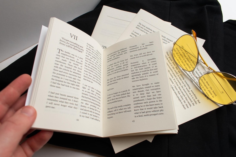 May include: An open book with text on its pages, surrounded by loose sheets of paper and yellow-tinted sunglasses. The book is held open by a hand, with the text visible. The sunglasses have a gold frame.