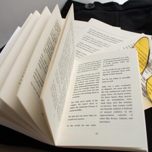 May include: An open book with pages fanned out, displaying printed text. A pair of round yellow-tinted sunglasses rests on a stack of loose papers. The items are placed on a black surface, suggesting a literary or intellectual theme.