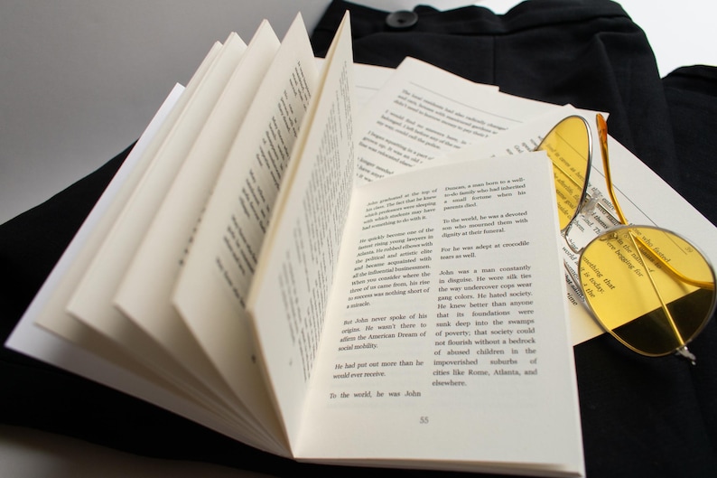 May include: An open book with pages fanned out, displaying printed text. A pair of round yellow-tinted sunglasses rests on a stack of loose papers. The items are placed on a black surface, suggesting a literary or intellectual theme.