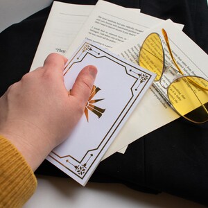 May include: A white card with a gold geometric design and a gold sunburst emblem is held over a stack of papers and a pair of yellow-tinted glasses. The glasses have a thin, gold frame. The items are on a black surface.