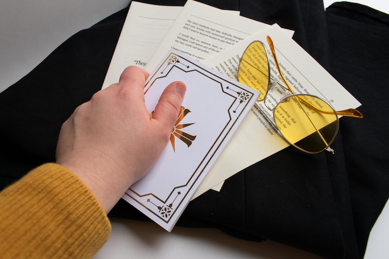 May include: A white card with a gold geometric design and a gold sunburst emblem is held over a stack of papers and a pair of yellow-tinted glasses. The glasses have a thin, gold frame. The items are on a black surface.