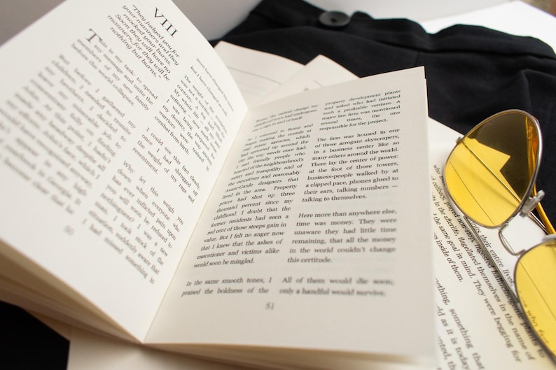May include: Open book with printed text, pages fanned out. Yellow-tinted aviator sunglasses rest on the right side. A black garment is visible in the background.