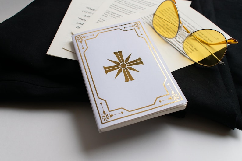 May include: A white book with a gold cross design and gold border, resting on a black surface. Yellow-tinted sunglasses and a stack of papers are also visible. The book's design is the focal point.