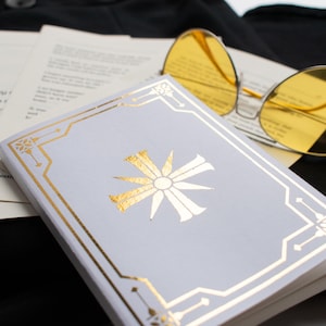 May include: A small, light gray book with gold accents and a gold sunburst design. Yellow-tinted sunglasses rest on top of the book and pages of text. The items are on a black surface.